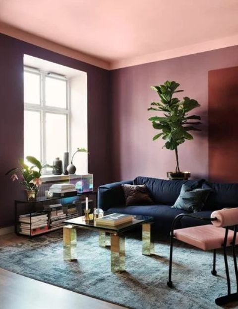 a colorful living room with a pink ceiling, purple walls, a navy sofa, a pink chair, a glass coffee table, a console and potted plants