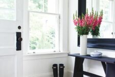 a cool entryway with a black frame mirror, a console table, white walls and a black floor, a jute rug and bold blooms