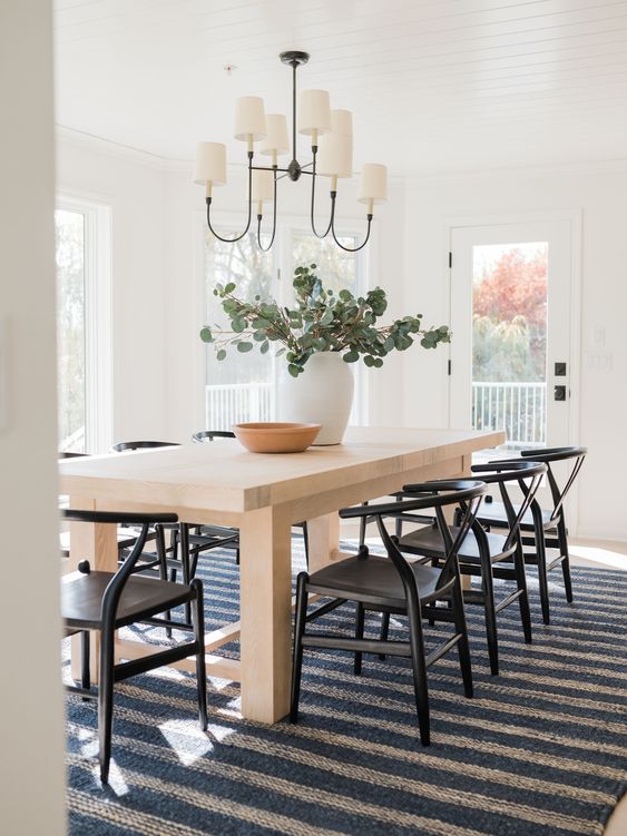A cool modern farmhouse dining room with a light stained table, black wishbone chairs, a chandelier and a striped rug
