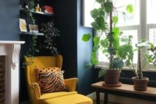 a cool nook by the window with navy walls, a bench with plants, built-in shelves with plants, a yellow Strandmon chair with an ottoman and a fireplace