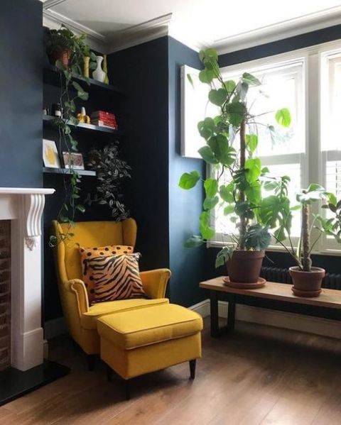 A cool nook by the window with navy walls, a bench with plants, built in shelves with plants, a yellow Strandmon chair with an ottoman and a fireplace