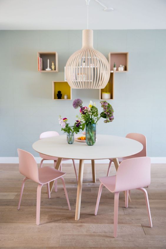a delicate and subtle dining space with sage green walls, a round table, pink chairs, a pendant lamp and box shelves