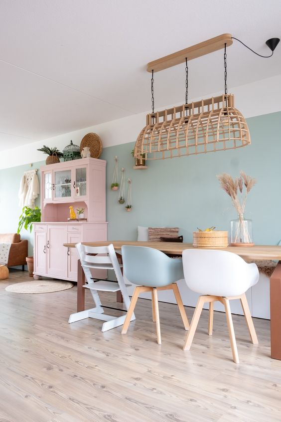 a delicate pastel dining room with sage green walls, a stained table, white and sage chairs, a woven pendant lamp and a pink buffet