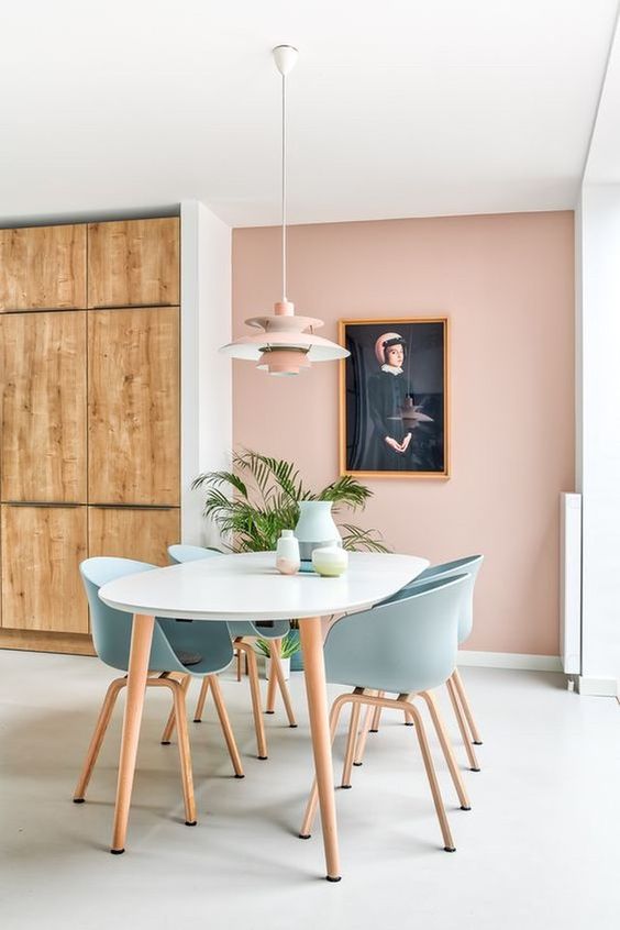 a dusty pink accent wall, a white dining table, pastel blue chairs, a pink pendant lamp, an artwork and a stained storage unit