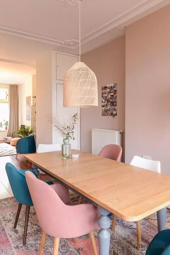 a dusty pink dining room with a stained table, dusty pink, white and teal chairs, a pink rug and a woven pendant lamp
