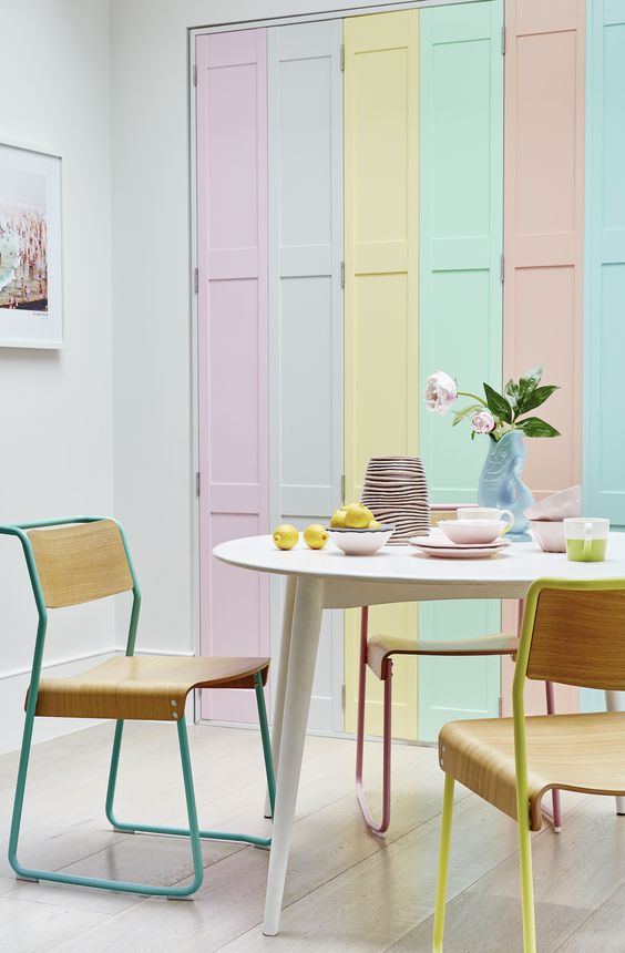 a fun pastel dining space with bright pastel shutters, an oval table, plywood chairs, pastel tableware and some blooms