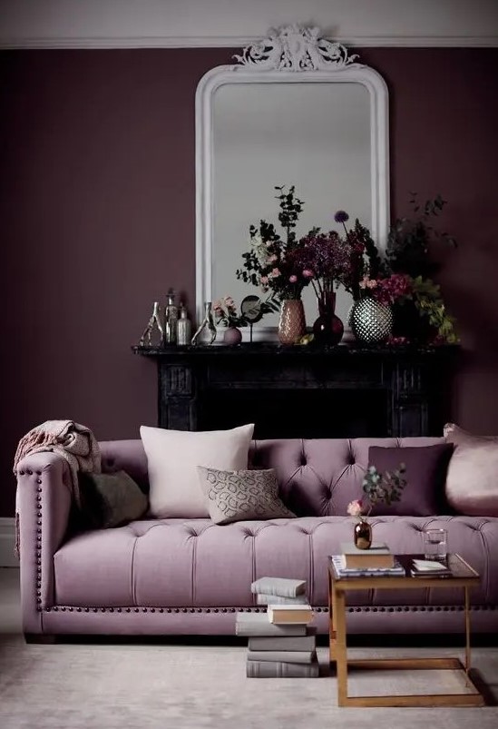 a gorgeous living room with a deep purple accent wall, a lilac sofa, a glass coffee table, a black fireplace and beautiful blooms on the mantel