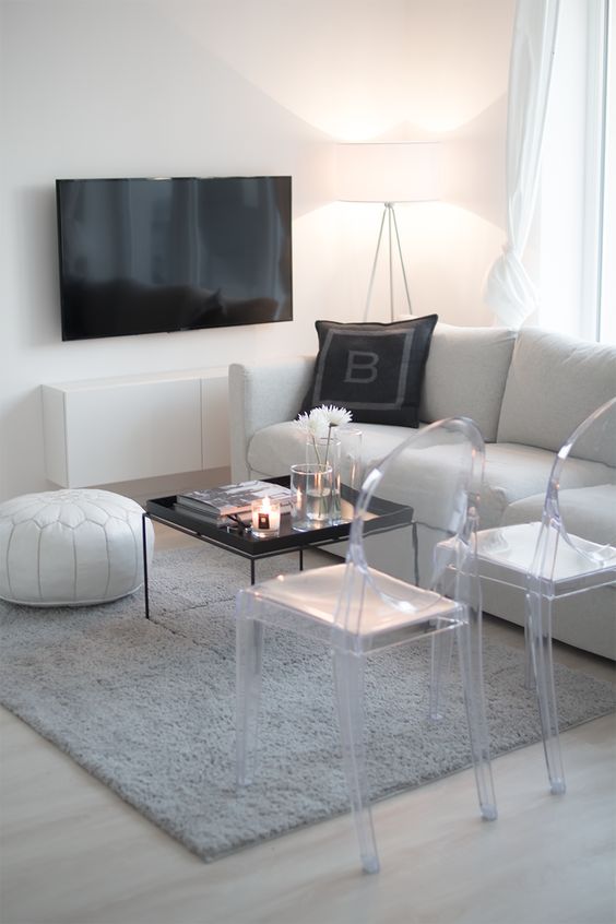 a laconic white living room with a sofa, a leather pouf, ghost chairs, a grey rug, a TV and a wall-mounted TV unit