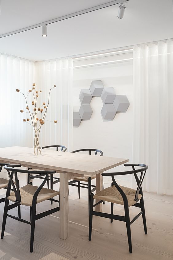 A light colored modern dining space with a light stained table, black wishbone chairs, dried blooms, hexagon panels