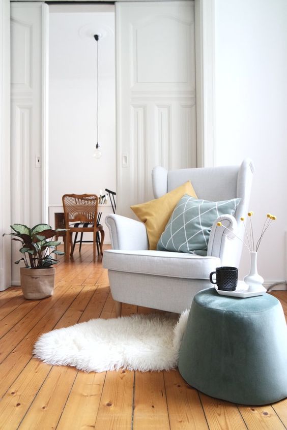 A light filled space with a Strandmon chair, a pale blue pouf, pillows, a faux fur rug and a potted plant is amazing