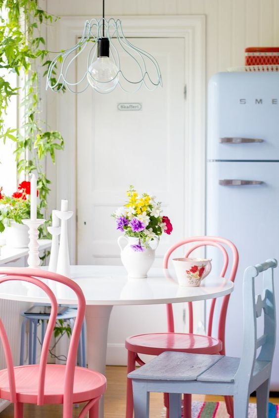a lovely dining nook with a round table, pink and a pale blue chair, a pale blue Smeg fridge and a pendant lamp with a blue wire lampshade