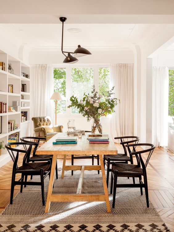a mid-century modern dining room with a light-stained table, black wishbone chairs, a black chandelier and a boho rug