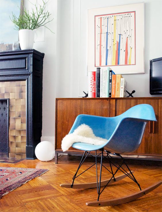 A mid century modern nook with a stained credenza, a blue Eames rocking chair, a non working fireplace, bold decor and books
