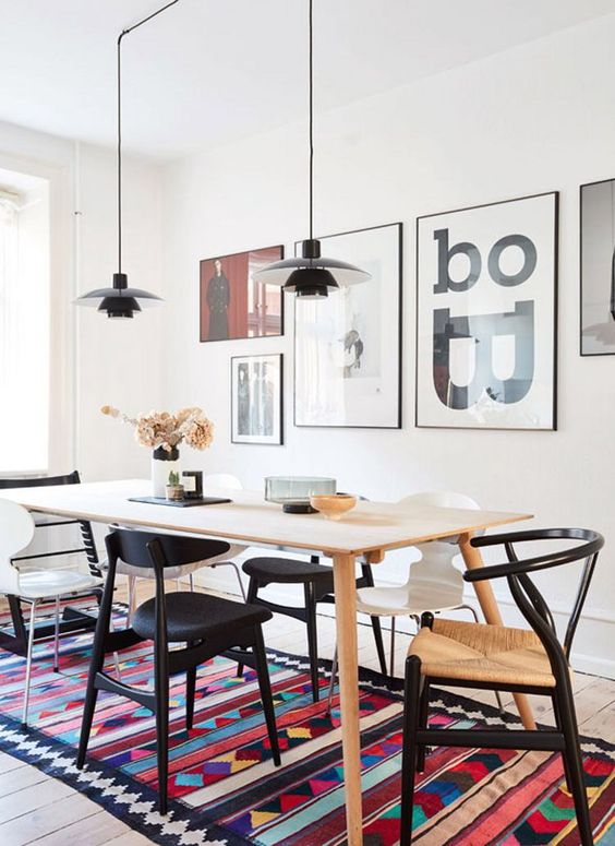 A modern boho dining space with a light stained table, black and white chairs including wishbone ones, a bold rug, a gallery wall