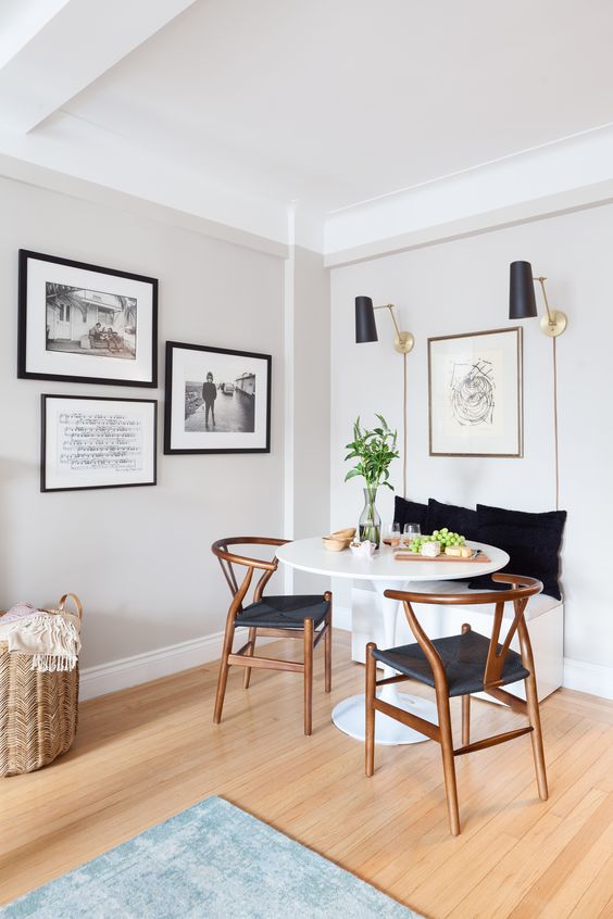 A modern dining nook with a built in bench, a round table, stained wishbone chairs, black sconces and artworks