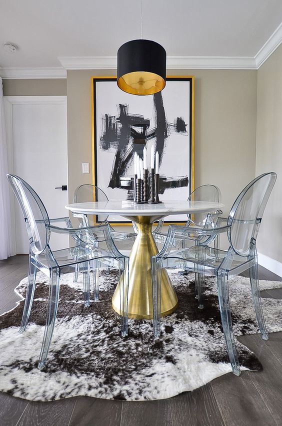 a modern refined dining space with greige walls, a round table with a gold base, ghost chairs, a bold artwork and a black pendant lamp