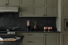 a moody olive green kitchen with shaker cabinets, a black tile backsplash and black stone countertops, gold touches