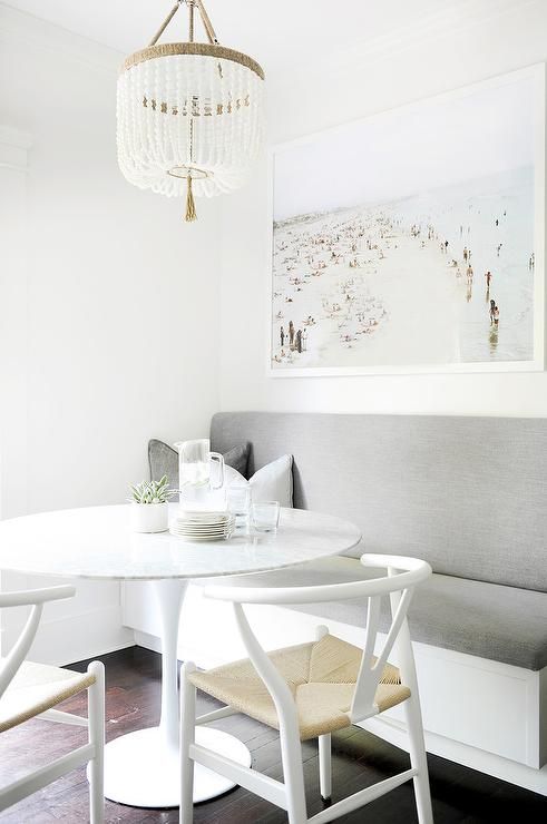 A neutral coastal dining room with a built in upholstered bench, a round table, white wishbone chairs, a large coastal artwork