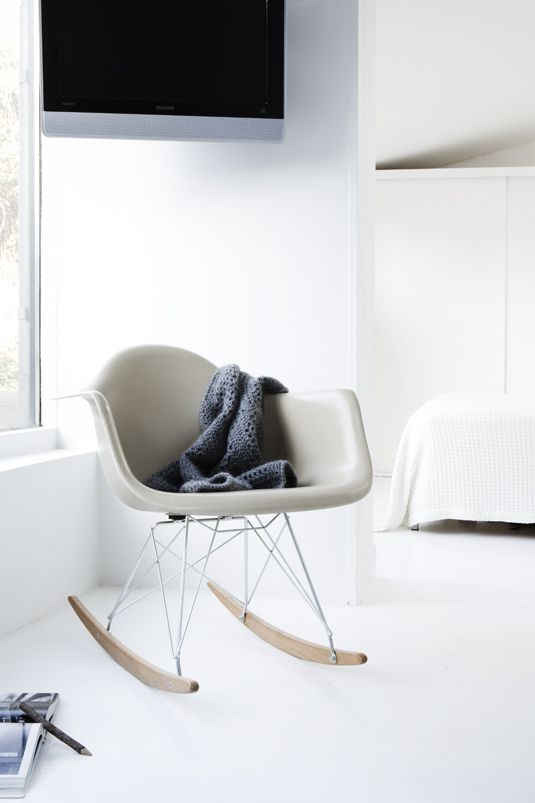 A neutral light filled space with a grey Eames rocker, a white bed and bedding, a built in cabinet for storage