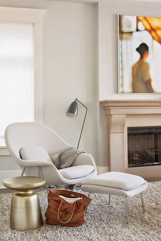 A neutral space with a built in fireplace, a neutral Wumb chair and ottoman, a polished metal side table and a floor lamp
