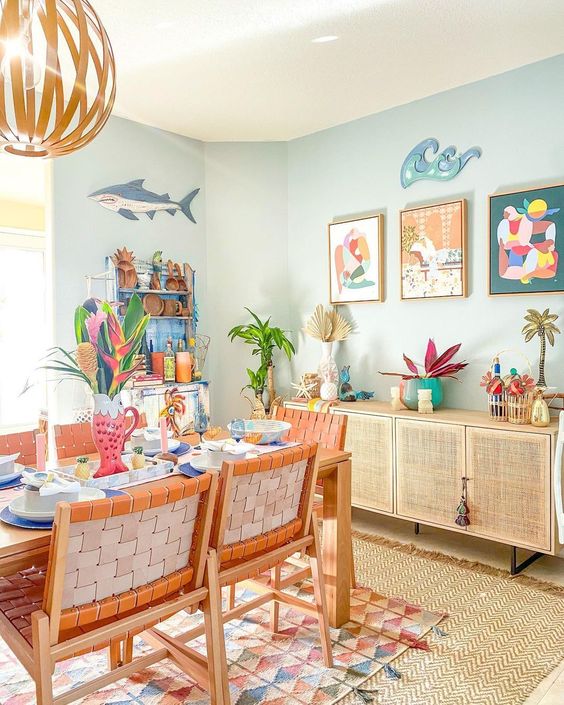 a pastel blue dining room with a rattan credenza, a stained table and orange chairs, layered rugs and colorful artwork