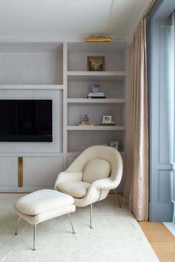 A peaceful neutral space with built in shelves, a creamy Wumb chair and ottoman, neutral curtains and brass details