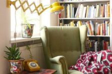 a retro cozy reading nook with a large bookcase, a green IKEA Strandmon wingback chair and a yellow wall lamp is very comfortable