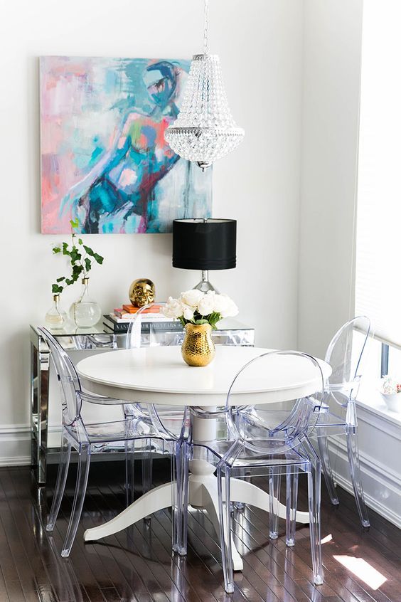 a small yet chic dining space by the window, with a round table, ghost chairs, a mirror console table, a bold artwork and a crystal chandelier