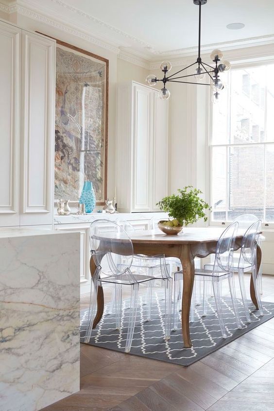a sophisticated dining room with creamy cabinetry, a stained dining table, ghost chairs, a printed rug and a marble slab console