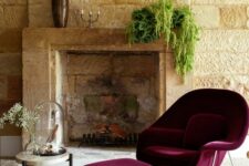 a sophisticated living room with vintage stone cladding, a mantel with greenery, a burgundy Wumb chair with an ottoman and a side table
