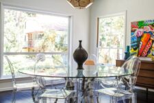 a sunroom as a dining room with a stained sideboard, a glass dining table with a unique base, ghost chairs and a faceted pendant lamp