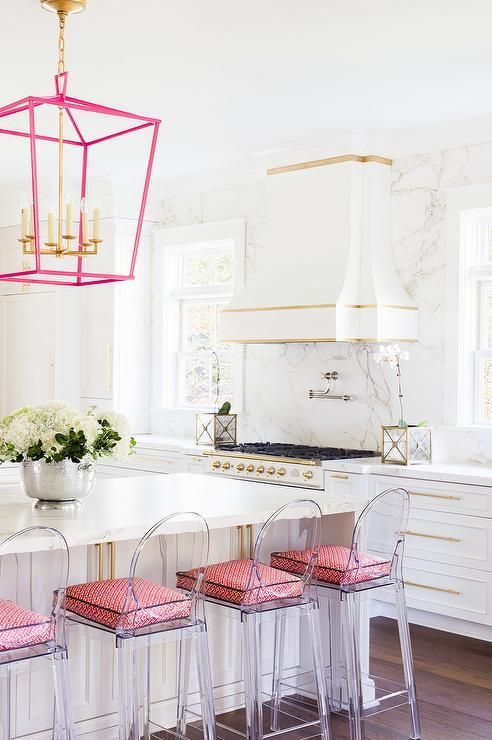 a white kitchen with shaker cabinets, a white marble backsplash and countertops, a kitchen island, ghost chairs with pink cushions and a pink frame pendant lamp
