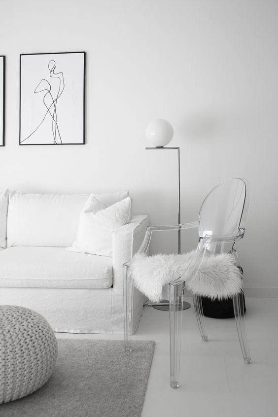 a white living room with a sofa, a pouf, a ghost chair with faux fur, a floor lamp and some black and white prints