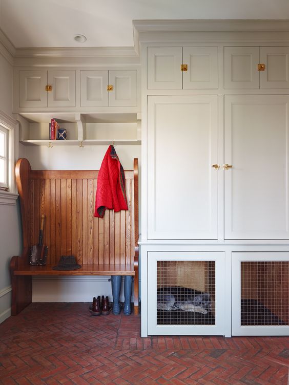 A white mudroom with a brick floor, creamy cabinets, a stained rack and a built in dog crate in the lower part