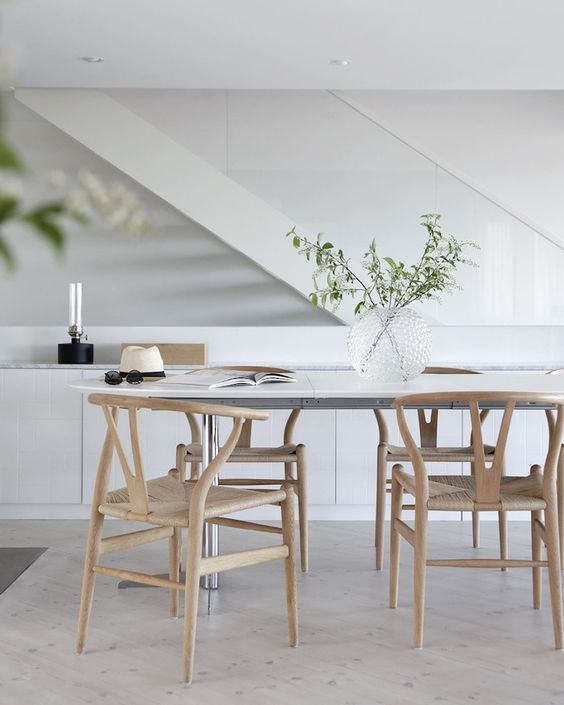 An airy Scandinavian dining space with a white dining table, light stained wishbone chairs, a sleek storage unit and greenery