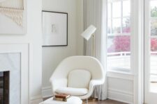 an airy space with a marble clad fireplace, a creamy chair with an ottoman, a floor lamp and artwork