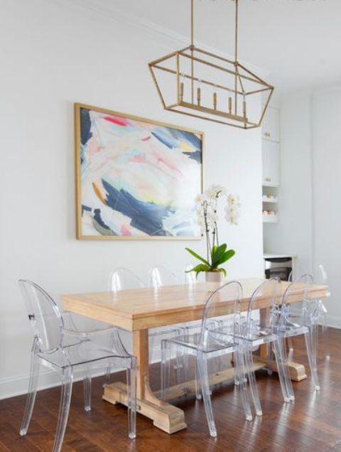An eclectic dining room with a light stained table, ghost chairs, a bold artwork and a gilded pendant lamp