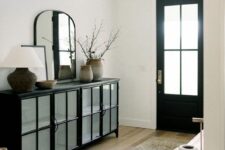 an elegant black and white entryway with a black door and a credenza, an arched mirror, a gold lantern and a white vase