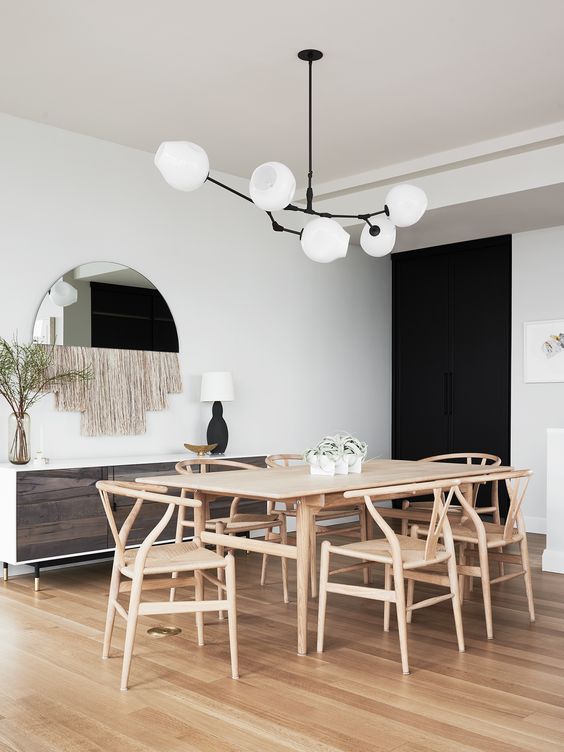 an elegant mid-century modern dining room with a light-stained table and wishbone chairs, a chandelier, a reclaimed wood credenza and a mirror with fringe