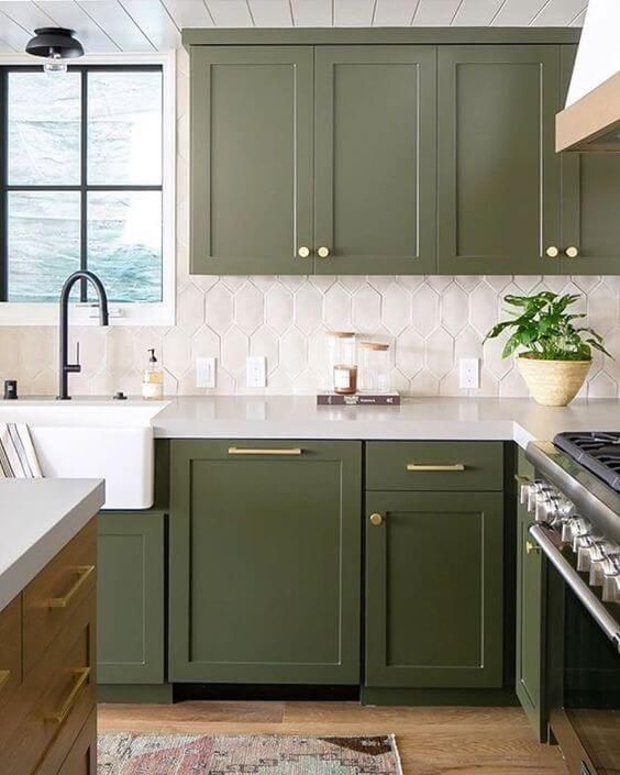 an olive green farmhouse kitchen with shaker cabinets, a catchy white tile backsplash, black fixtures and gold handles and knobs