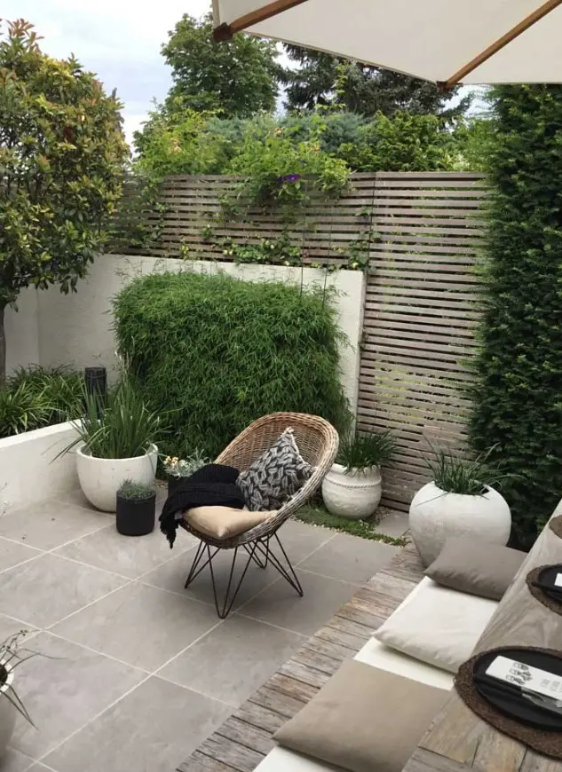 a contemporary walled backyard with a planked screen, with a tiled floor, potted grasses and greenery, a small dining set with pillows and a woven chair
