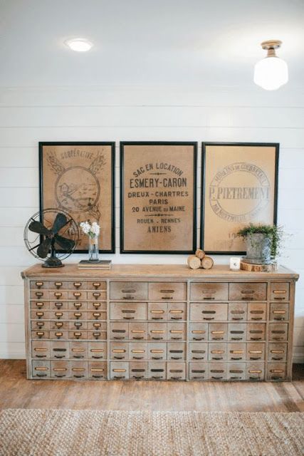 a light-stained card cabinet is a beautiful vintage storage unit, rustic vintage art over it and greenery in a bucket add interest to it