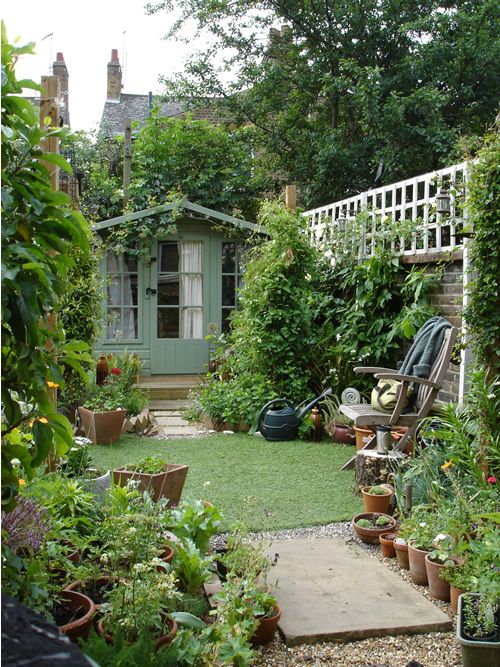 a small and narrow garden with a green lawn, greenery and shrubs, potted plants and a chair with a side table
