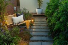 20 a long and narrow garden with a tile pathway, greenery, grasses and blooms, a rattan bench with pillows and some lights
