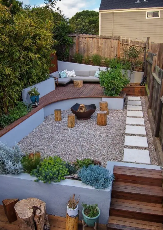 a small and welcoming backyard with a fire pit zone and tree stumps with growing plants and trees, with a raised sitting zone with a sofa