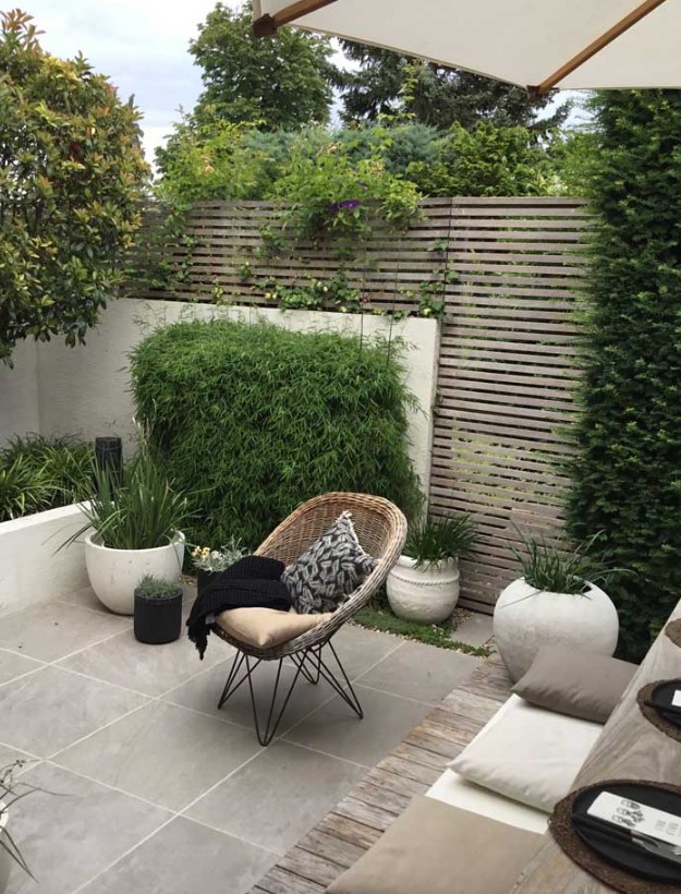 a modern paved patio with a planked screen, with a tiled floor, potted grasses and greenery, a small dining set with pillows and a woven chair