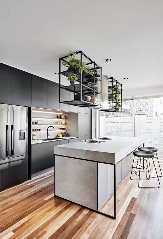 A modern industrial kitchen in black, with built in shelves, a concrete kitchen island, suspended shelves over it and black stools
