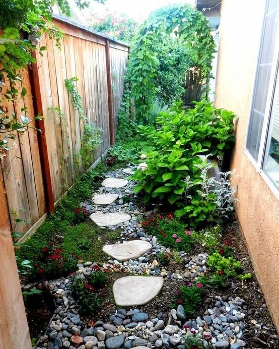 a small and bright side garden nook with pebbles, greenery and bright blooms, shrubs and a climbing plants on the fence