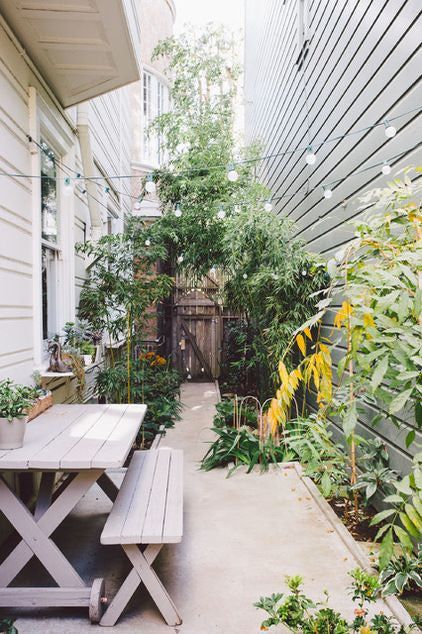 a small side garden space clad with concrete, wiht greenery and plants, with a whitewashed dining set and lights over the space