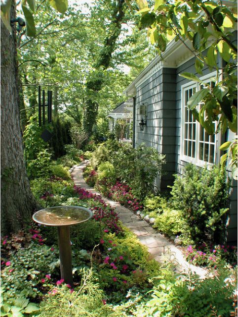 a little and lovely side garden with lots of greenery and blooms, with a stone pathway and a bird bath plus some trees